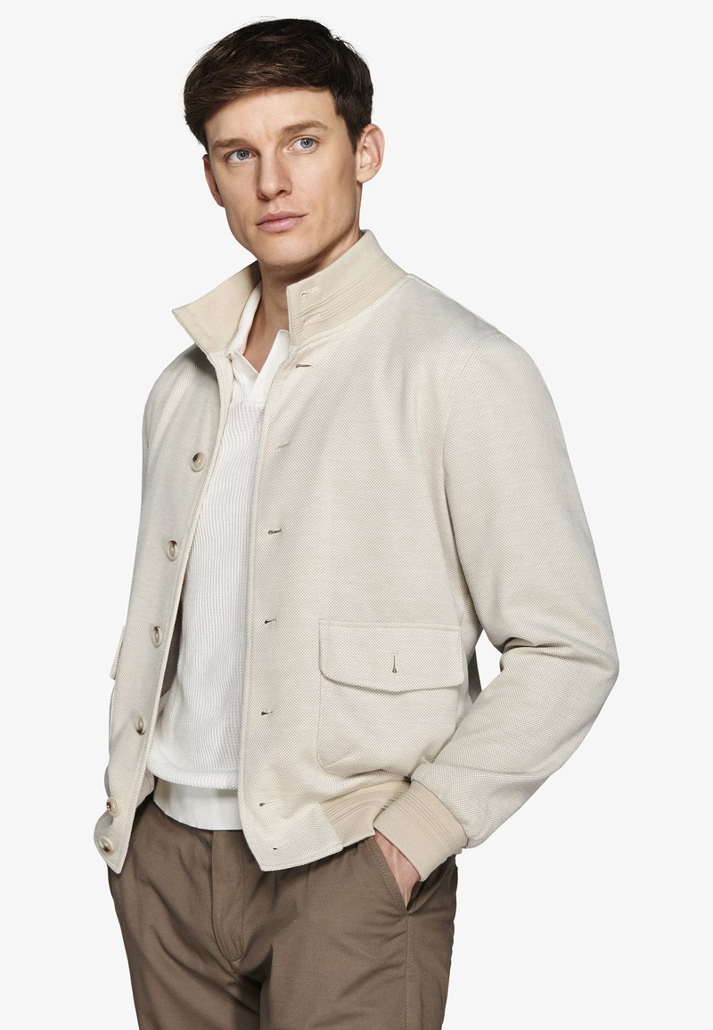 Mand iført en beige jakke med knapper over en hvid sweater og brune bukser, der kigger let til siden med hænderne i lommerne.