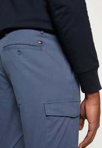 Graue Cargo-Hosen mit glatter Textur, ausgestattet mit einer Gesäßtasche, Knopfverschluss und einem kleinen Logo in Rot, Weiß und Blau auf der Tasche.