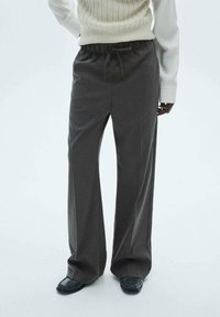 Pantalon gris à jambes larges en tissu texturé, avec une taille élastique et un cordon de serrage. Associé à des chaussures noires et brillantes.