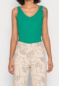 Zelený žebrovaný top bez rukávů s výstřihem do V, doplněný béžovými kalhotami se vzorem ručně nakreslených rukou v černé barvě s Tyrkysovými akcenty.