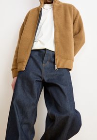 Tan ribbad zip-up jacka över en vit skjorta, matchad med mörkblå vida denimbyxor med gul sömnad och en dragkedjeförsedd detalj.