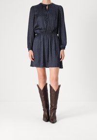 Nainen, jolla on yllään tummansininen pitkähihaista mekkoa, jossa on pieniä valkoisia ja punaisia pisteitä, yhdistettynä korkeisiin ruskeisiin nahkaiseen cowboy-kenkään.