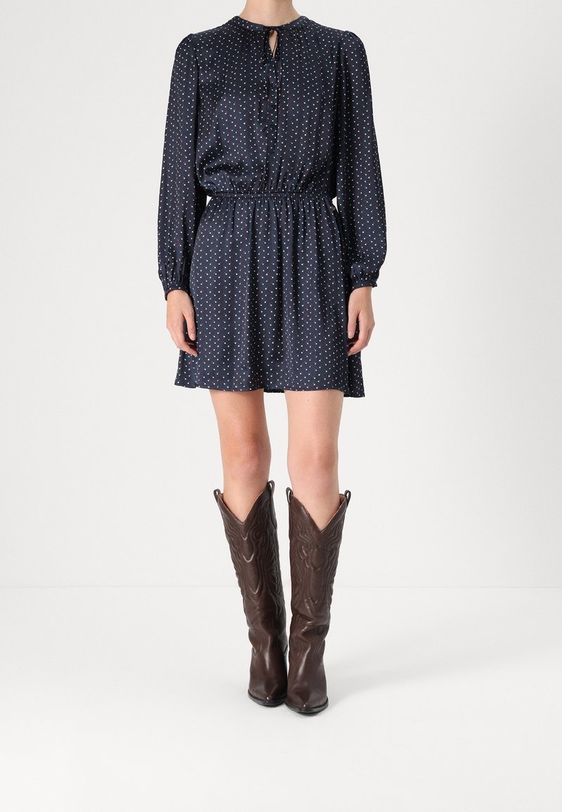 Nainen, jolla on yllään tummansininen pitkähihaista mekkoa, jossa on pieniä valkoisia ja punaisia pisteitä, yhdistettynä korkeisiin ruskeisiin nahkaiseen cowboy-kenkään.