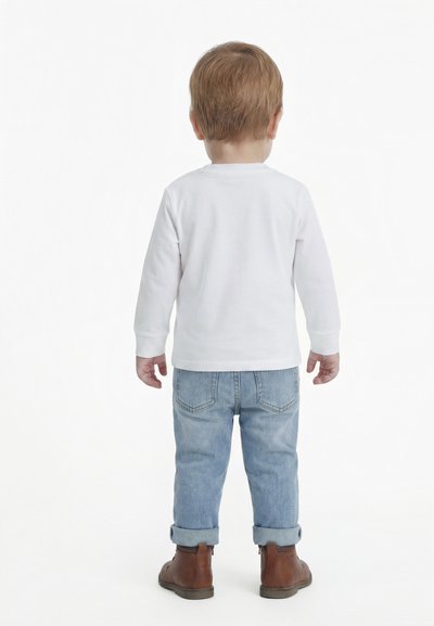 Niño pequeño con cabello castaño claro visto de espaldas, vestido con una camiseta de manga larga blanca, jeans azul claro remangados y botas marrones.