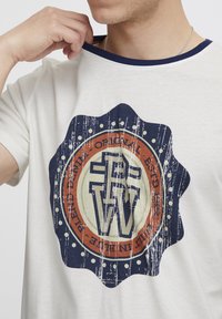 Kremfarget t-skjorte med korte ermer som har en sirkulær logo i blå, oransje og hvite farger. Designet har en slitt tekstur og prikkete detaljer.