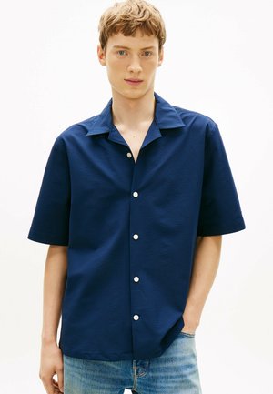 Junger Mann, der ein strukturiertes dunkelblaues Hemd mit kurzen Ärmeln und weißen Knöpfen sowie hellblaue Jeans trägt, die Hand in der Hosentasche.