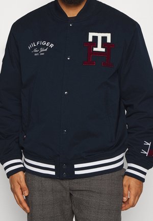 Hombre con chaqueta abotonada azul marino con logo "TH" en blanco y burdeos, texto "Hilfiger New York EST. 1985" y puños a rayas.