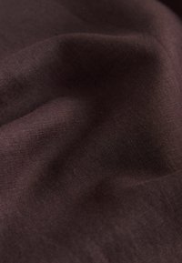 Material moale, pliat, de culoare maro închis, cu textură fină și o strălucire subtilă, prezentând curbe line și detalii fine ale țesăturii.