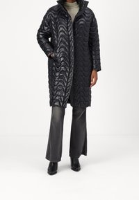 Abrigo negro largo acolchado con patrón de ondas, con cuello alto y bolsillos laterales. Combinado con pantalones grises de pierna ancha y botines negros.