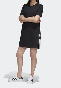 Kvinna iförd svart Adidas klänning med kort ärm och slits på sidan, samt vita Adidas sneakers med svarta ränder, står med armarna i kors.