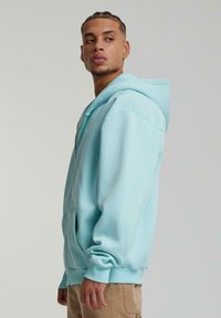 Lyseblå zip-up hoodie lavet af blød bomuldsblanding, der har en rummelig hætte, lange ærmer og en front kangaroo lomme.