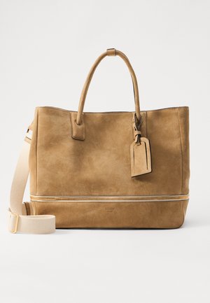 Sac fourre-tout en suède tan avec double poignées, une bandoulière amovible, un compartiment inférieur zippé et une étiquette de bagage attachée.