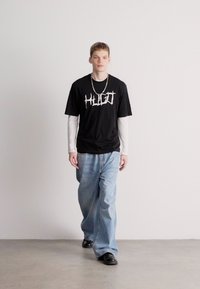 Μαύρη μπλούζα με λευκό γραφικό κείμενο, φορεμένη πάνω από ένα λευκό πουκάμισο με μακριά μανίκια, σε συνδυασμό με χαλαρές ανοιχτό μπλε τζιν και μαύρα παπούτσια.