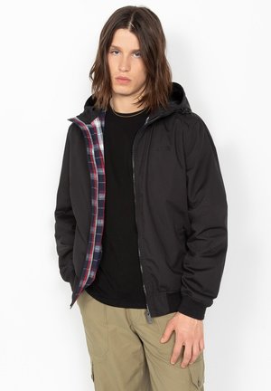 Jeune homme portant une veste à capuche noire avec doublure à carreaux, une chemise noire et un pantalon cargo beige, debout les mains dans les poches.