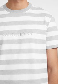 Hall värvi ja valge triibuline T-särk ümarate kaelustega, mille keskosas on reljeefne 'GUESS JEANS' logo. Puuvillane kangas, lühikesed varrukad.