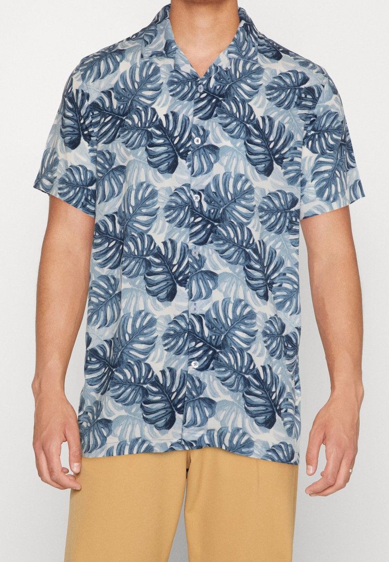 Camicia a maniche corte con un motivo di foglie tropicali blu e verdi su uno sfondo chiaro. Presenta un colletto e una chiusura frontale con bottoni.