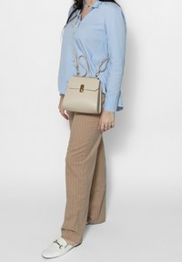 Borsa a tracolla beige con design strutturato, chiusura in hardware dorato, tracolla regolabile e una superficie liscia, indossata con camicia blu e pantaloni a righe.