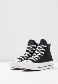 Baskets montantes noires en toile avec des lacets blancs, une semelle en caoutchouc texturée et une base blanche avec une bande noire.