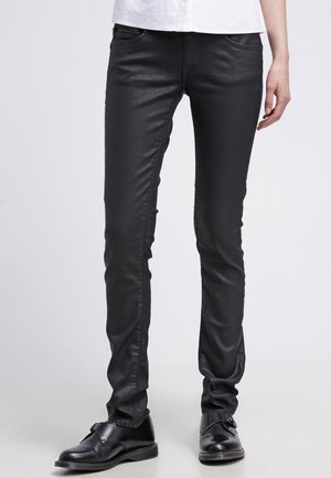 Jeans negros ajustados hechos de un material liso y brillante, con bolsillos frontales y un corte estrecho, combinados con zapatos de cuero negros.