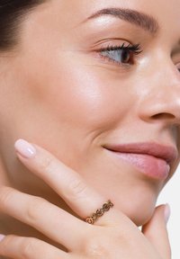 Anello in oro rosa con un design texturizzato che presenta piccole aperture circolari, indossato su una mano con unghie rosa pallido, su uno sfondo neutro.