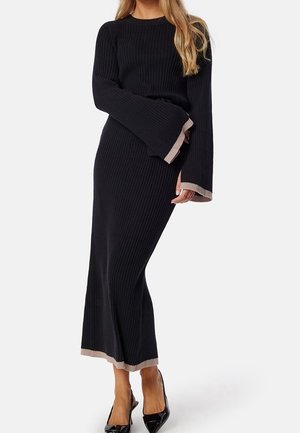 Femme portant une robe noire en maille côtelée avec des manches évasées et une bordure beige, associée à des escarpins noirs à bout pointu.