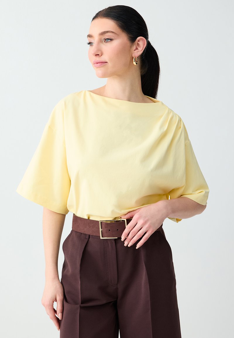 Femme aux cheveux noirs attachés en queue de cheval basse, portant un haut ample jaune pâle rentré dans un pantalon taille haute marron foncé avec une ceinture large.