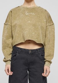 Olivově zelený zkrácený sweatshirt s texturovaným povrchem a bílou grafikou. V kombinaci s černými džíny s vysokým pasem, které mají zapínání na knoflík.