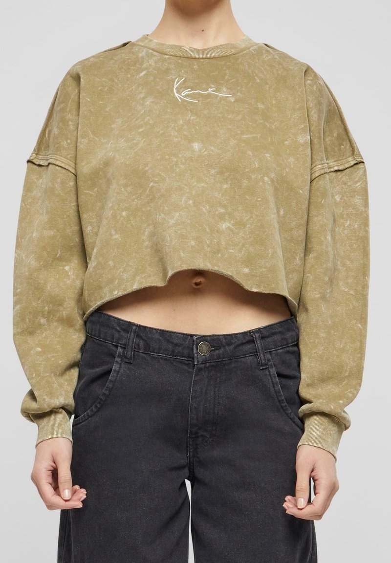 Olivově zelený zkrácený sweatshirt s texturovaným povrchem a bílou grafikou. V kombinaci s černými džíny s vysokým pasem, které mají zapínání na knoflík.