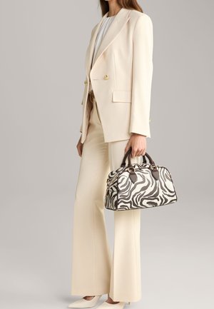 Donna in completo crema che tiene una borsa con stampa zebrata bianca e nera con manici scuri, in piedi contro uno sfondo chiaro e uniforme.