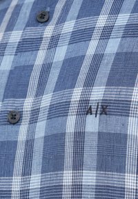 Μπλε και λευκό καρό πουκάμισο με μαύρα κουμπιά και μικρή μαύρη κεντημένη «A|X» στην περιοχή του στήθους.