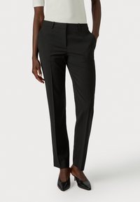 Woman wearing slim black tailored trousers and black pointed-toe heels, with hands in pockets, standing against white background.