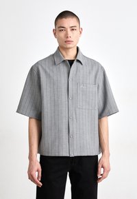 Cămașă scurtă cu mânecă, gri cu dungi, cu guler, buzunar frontal și închidere cu nasturi. Materialul pare neted, cu un model subtil. Se poartă pantaloni negri.