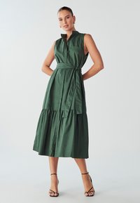 Roheline, ilma varrukateta kleit, millel on nupud ees, vöödiga talje ja kihtide äär. Valmistatud siledast puuvillast kangast, on sellel rinna tasku.