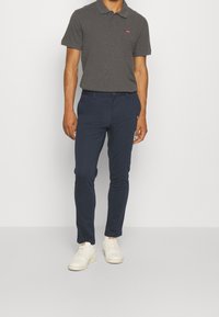 Polo gris foncé avec deux boutons et logo rouge, associé à un pantalon slim-fit bleu marine et des baskets blanches, se tenant devant un fond uni.
