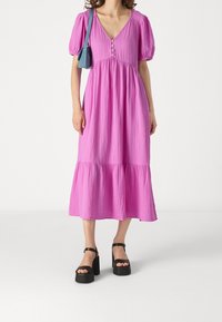 Rosa midiklänning med V-hals, puffärmar, knapper och lager på lager-hamn. Kombinerad med svarta platåsandaler för kontrast.