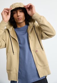 Jovem com cabelo encaracolado ajeita o capuz de um casaco bege com fecho, sobre uma camisa azul, posando contra um fundo claro e simples.