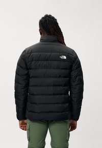 Chaqueta acolchada negra con diseño de acolchado, con un alto cuello y un pequeño logo blanco en el hombro izquierdo. Material suave y acolchado.