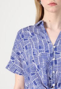 Korte ermer bluse med et blått og hvitt abstrakt bølgedesign, med krage og knappelukking. Myk stoffkvalitet.