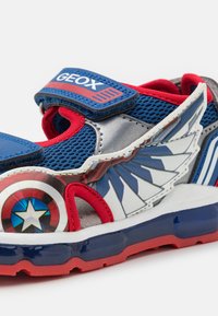 Blå og sølv atletiske sko med røde accenter, der har netmateriale, et velcrobånd og et grafisk design af et skjold og vinger.