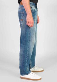 Lichtblauwe denim jeans met een rechte snit, voorzien van achterzakken, koperen klinknagels en een versleten textuur. Draagt met witte sneakers.