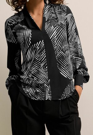 Vrouw draagt een zwart-wit blouse met een geometrisch patroon, lange mouwen en zwarte broek, handen in de zakken, staand tegen een effen achtergrond.
