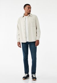 Ljusbeige jacka med krage och framficka, bärs över en vit skjorta och blå jeans, i kombination med svarta sneakers med vita detaljer.