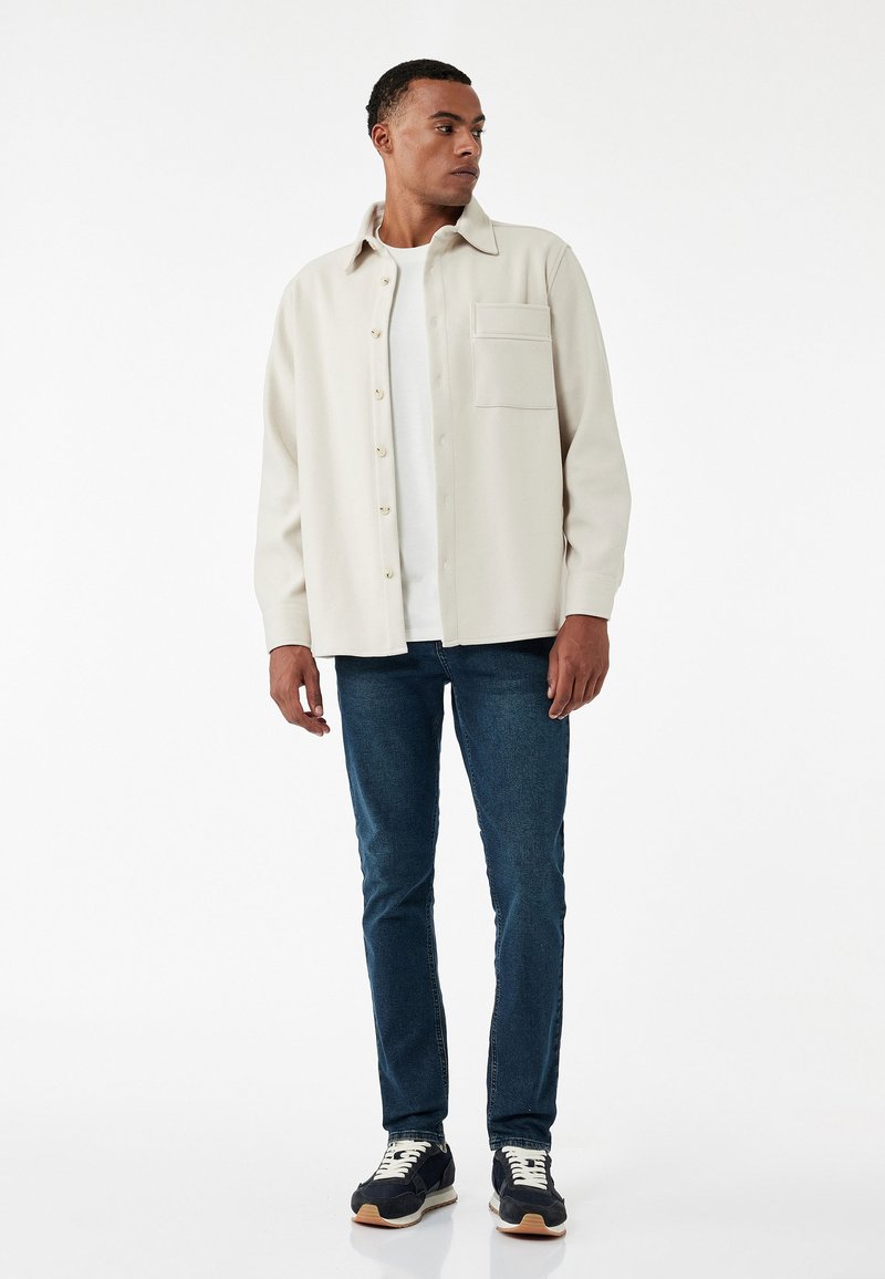 Ljusbeige jacka med krage och framficka, bärs över en vit skjorta och blå jeans, i kombination med svarta sneakers med vita detaljer.