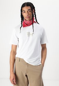 Tricou alb cu mânecă scurtă, care prezintă un text grafic aurit "THE PLANT" pe piept, combinat cu pantaloni bej și o bentiță roșie.