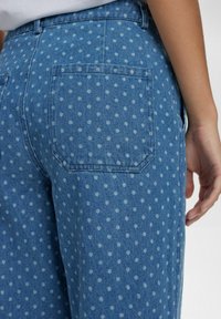 Mėlyni džinsai su taškeliais, turintys nugarinę kišenę, aukštu liemeniu ir laisvu siluetu. Audinys turi minkštą tekstūrą.