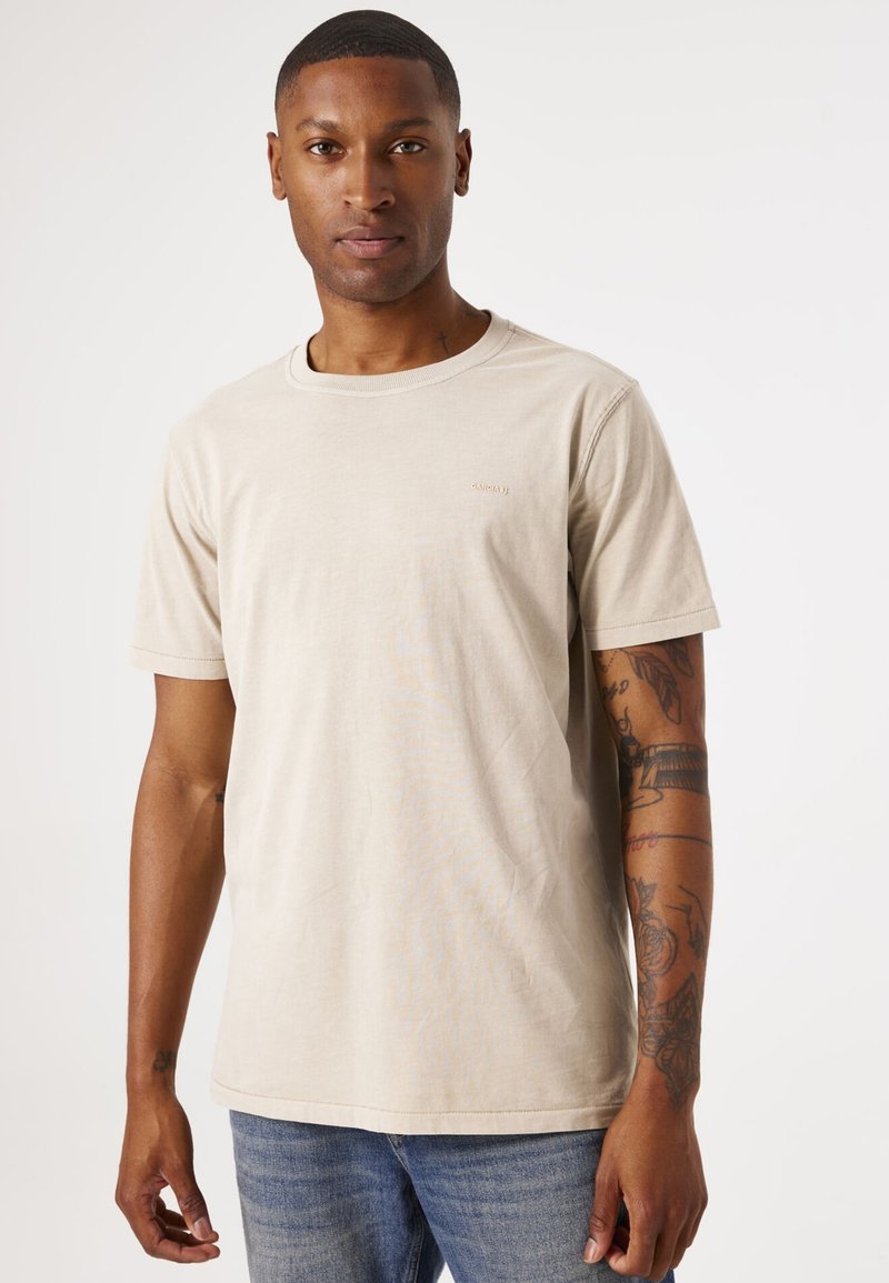 Helle beige Baumwoll-T-Shirt mit Rundhalsausschnitt und kurzen Ärmeln. Mit kleinem Logo auf der Brust. Kombiniert mit blauen Jeans.