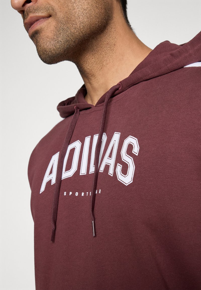 Hoodie bordeaux avec cordon de serrage, comportant un logo "ADIDAS" blanc et du texte "SPORTSTAR". Le tissu semble doux avec une texture lisse.