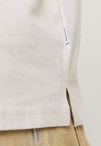 Lys beige bomull T-skjorte med sideslitser og en liten etikett ved kanten. Myk tekstur, avslappet design og rett kutt.