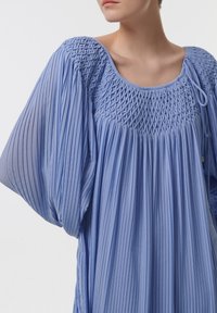 Vestido plisado de un azul claro, con cuerpo fruncido, mangas anchas y textura sutil. Degradado de tonos más oscuros a más claros en los bordes.