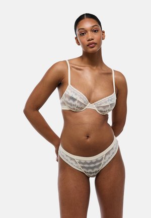 Femme portant un soutien-gorge en dentelle blanche transparente et une culotte assortie, debout les mains derrière le dos sur un fond uni.